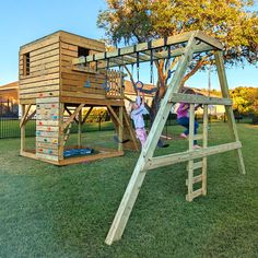 two children playing on a wooden play set in the grass with a tree house behind them