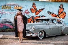 a woman in a graduation cap and gown standing next to an old car with butterflies painted on the wall behind her