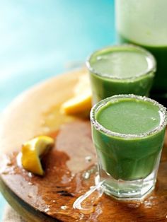 two glasses filled with green smoothie sitting on top of a wooden cutting board next to lemon wedges