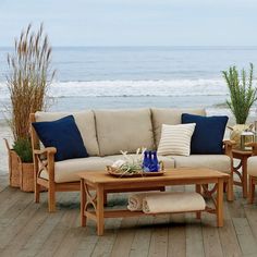 a couch and two chairs on a wooden deck near the ocean with plants in vases