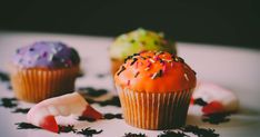 three cupcakes with sprinkles are on a white plate and one has orange frosting