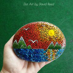 a hand holding a painted rock in front of a green background that says dot art by david reed