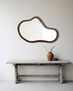 a wooden table with a vase and mirror on it next to a bench in front of a white wall