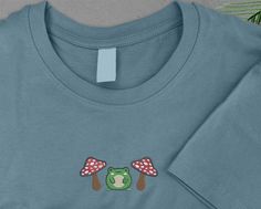 Mushroom Shirt, Adorable Embroidered Frog And Mushroom T-Shirt, Toadstool Woodland Aesthetic Cute Frog Shirt, Fairycore Mushroom Lover Gift PRODUCT SPECIFICATIONS: - Fabric: 100% Cotton. - Weight: White 193 gsm, Cols 203 gsm - Pre-shrunk. - Ribbed collar. - Taped neck and shoulders. - Twin needle sleeves and hem. - Tubular body. - Tear out label (some colours in transition). If you have any questions or query related to our Etsy listing, please ask through Etsy message center so we can assist an Fairycore Mushroom, Woodland Aesthetic, Embroidered Frog, Frog And Mushroom, Mushroom T Shirt, Frog Shirt, Message Center, Mushroom Shirt, Cute Frog