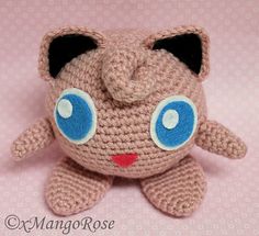 a crocheted stuffed animal with big blue eyes on a pink background that looks like a cat