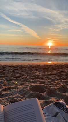 an open book sitting on top of a sandy beach next to the ocean at sunset