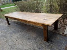 a large wooden table sitting on top of a cement ground next to bushes and trees