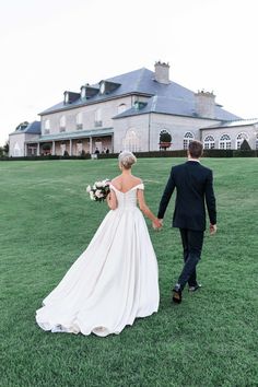 a bride and groom holding hands walking in front of a large house on the grass