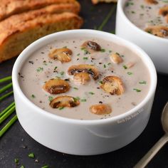 two white bowls filled with soup next to some bread and green chives on a black surface