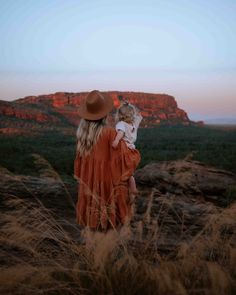 a woman in an orange dress and hat holding a small child on top of a hill