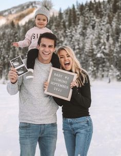 a man and woman holding a baby while standing in the snow with a sign that says baby 2 is coming together