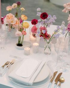 the table is set with white plates, silverware and colorful flowers in vases