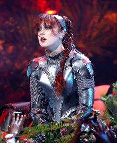 a woman with red hair wearing armor and gloves on her head, sitting in front of plants