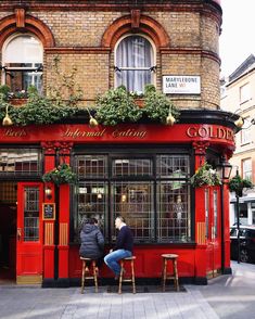 two people sitting at a red bar in front of a brick building with ivy growing on the windows