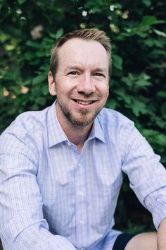 a man sitting on the ground in front of some bushes and trees smiling at the camera