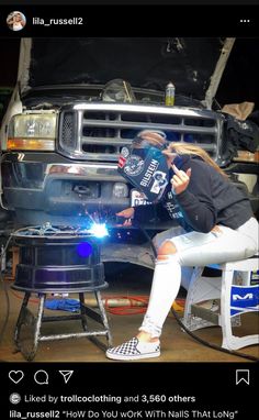 a woman sitting on a stool next to a grill in front of a truck with the hood up