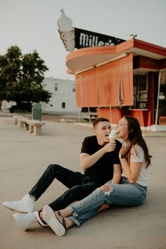 a man and woman sitting on the ground eating ice cream from a paper cup in front of a building