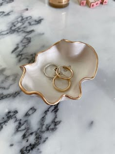 two gold wedding rings in a white bowl on a marble countertop next to lipstick