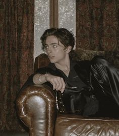 a young man sitting on top of a brown leather chair in front of a window