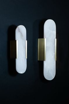 two white marble and brass sconces on a black wall, one is shaped like a rectangle