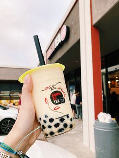 a person holding up a drink in front of a building with cars parked on the street