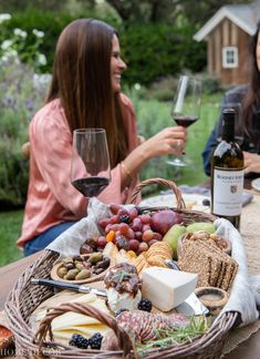 two women sitting at a table with wine and cheeses on it, while one woman holds a glass of wine