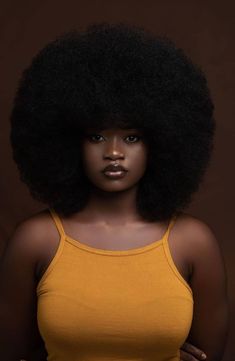 a woman with an afro standing in front of a brown background wearing a yellow tank top