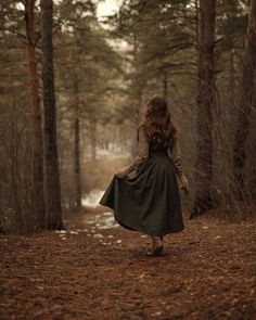 a woman is walking through the woods in a green dress and brown cardigan coat