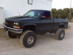 a black pickup truck parked in front of a white building with large tires on it