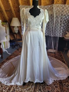 This is a Bridal Original from 1986, whose label was lost in the alteration process. The gown has been expertly hemmed and taken it, but the fabric remains with about an inch on each side that could be let out. Made from high-quality, mid-weight ivory Duchesse satin (non-silk) and imported French alencon lace that has been extensively pearled on the bodice and modified Juliet sleeves, which are gorgeous! It has a full aline skirt that lays close to the body due to inverted box pleats. Natural waist with tri-pleated attached belt, there is several inches of that inside the dress, as well. The neckline is a sweetheart shape that becomes an Elizabethan collar on the neck (so regal looking!) The back is covered and has a zipper closure. (I would consider adding pearl buttons for dramatic effec Cream Floor-length Gown With Lace Trim, Satin Wedding Gown With Lace Trim, Silk Wedding Gown With Long Train, Wedding Gown With Lace Bodice And Satin Material, Satin Wedding Gown With Lace Bodice, Debutante Ball Gown With Lace Trim And Fitted Bodice, Wedding Gown With Lace Bodice In Satin, Cream Satin Gown With Fitted Bodice, Formal Cream Satin Gown