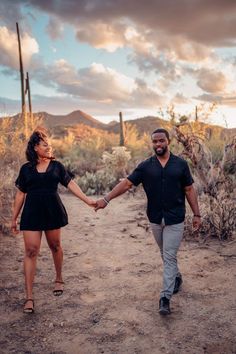 a man and woman holding hands while walking through the desert with cactus in the background