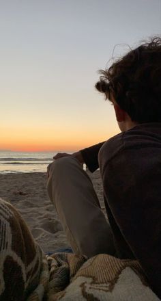 a man sitting on top of a beach under a blanket next to the ocean at sunset