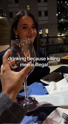 a woman sitting at a table with a wine glass in her hand and the words drinking because killing men is illegal