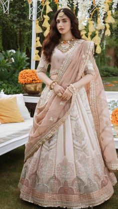 a woman in a white and pink lehenga standing on grass with an orange flower arrangement behind her