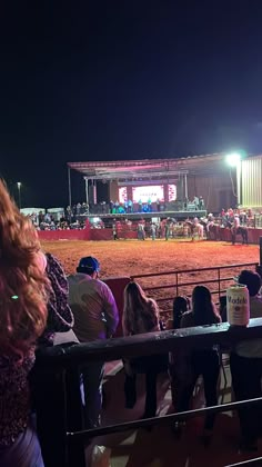 people are standing in front of a fence watching a horse race at night time with bright lights on the stands