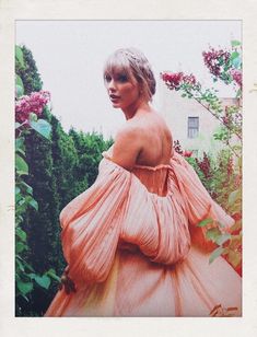 a woman in an off the shoulder dress standing next to some bushes and purple flowers