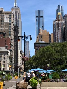 people are walking on the sidewalk in front of tall buildings and tables with umbrellas