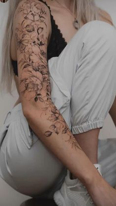 a woman sitting on the ground with her arm covered by flowers and leaves tattoo design