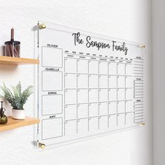 a wall mounted calendar on a whiteboard with wooden shelves and potted plants next to it