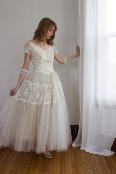 a woman in a white dress standing next to a window with her hands on the curtain