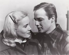 a man and woman standing next to each other in front of a clock tower, looking at each other