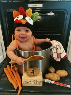 a baby wearing a turkey hat sitting in an oven with some carrots and potatoes