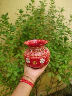 a hand holding a red and gold vase with flowers on the outside, in front of a bush