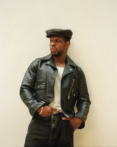 a man in a leather jacket and hat leaning against a wall with his hands on his hips