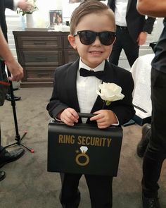 a little boy dressed in a tuxedo and holding a ring security box with a flower