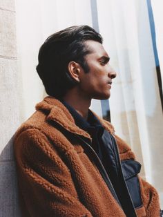 a man in a brown jacket leaning against a wall with his head turned to the side