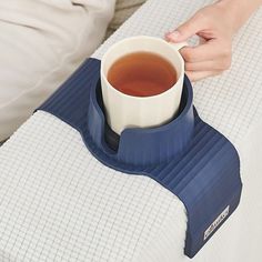 a person holding a cup of tea on top of a bed with their arm resting on the pillow