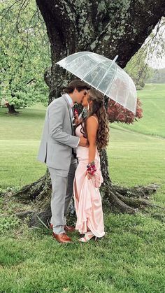 a bride and groom kissing under an umbrella in front of a tree on the grass