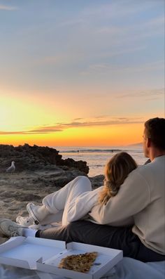 a man and woman sitting on the beach at sunset