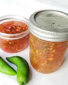 two jars filled with pickles next to green peppers on a white counter top,